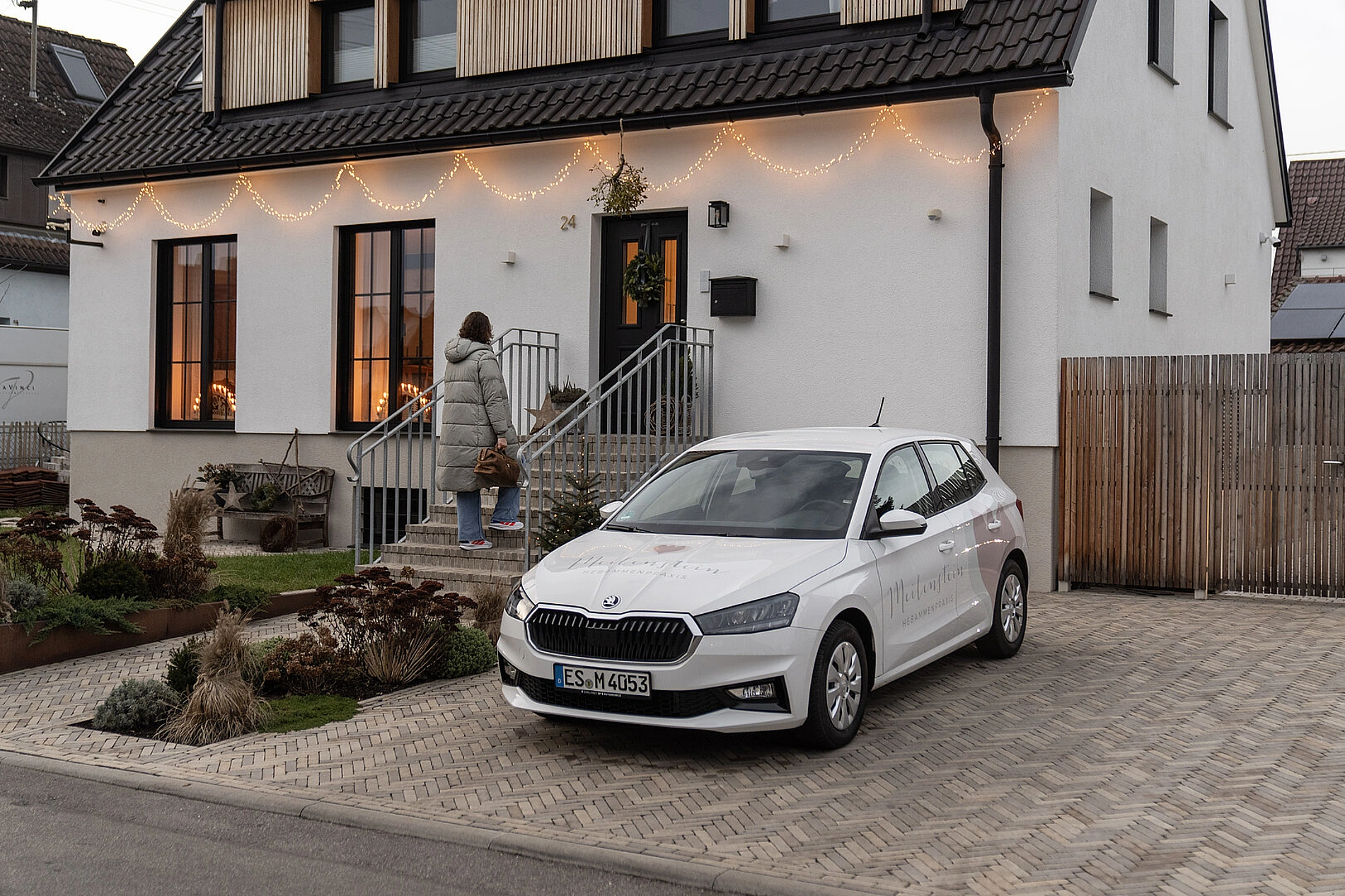 Das Bild zeigt eine Hebamme der Praxis Meilenstein während eines Hausbesuchs in Neuhausen auf den Fildern. Im Vordergrund steht ein weißes Auto in der gepflasterten Einfahrt eines modernen Einfamilienhauses. Die Fassade des Hauses ist warm beleuchtet, und eine Frau geht die Treppe zum Eingang hoch – symbolisch für den persönlichen, einfühlsamen und individuellen Betreuungsansatz der Hebammenpraxis. Das Bild vermittelt eine ruhige, vertrauensvolle Atmosphäre und unterstreicht den Fokus der Praxis Meilenstein auf mobile Wochenbettbetreuung, Hausbesuche und persönliche Betreuung von Familien in Neuhausen auf den Fildern und der Region Filderstadt.