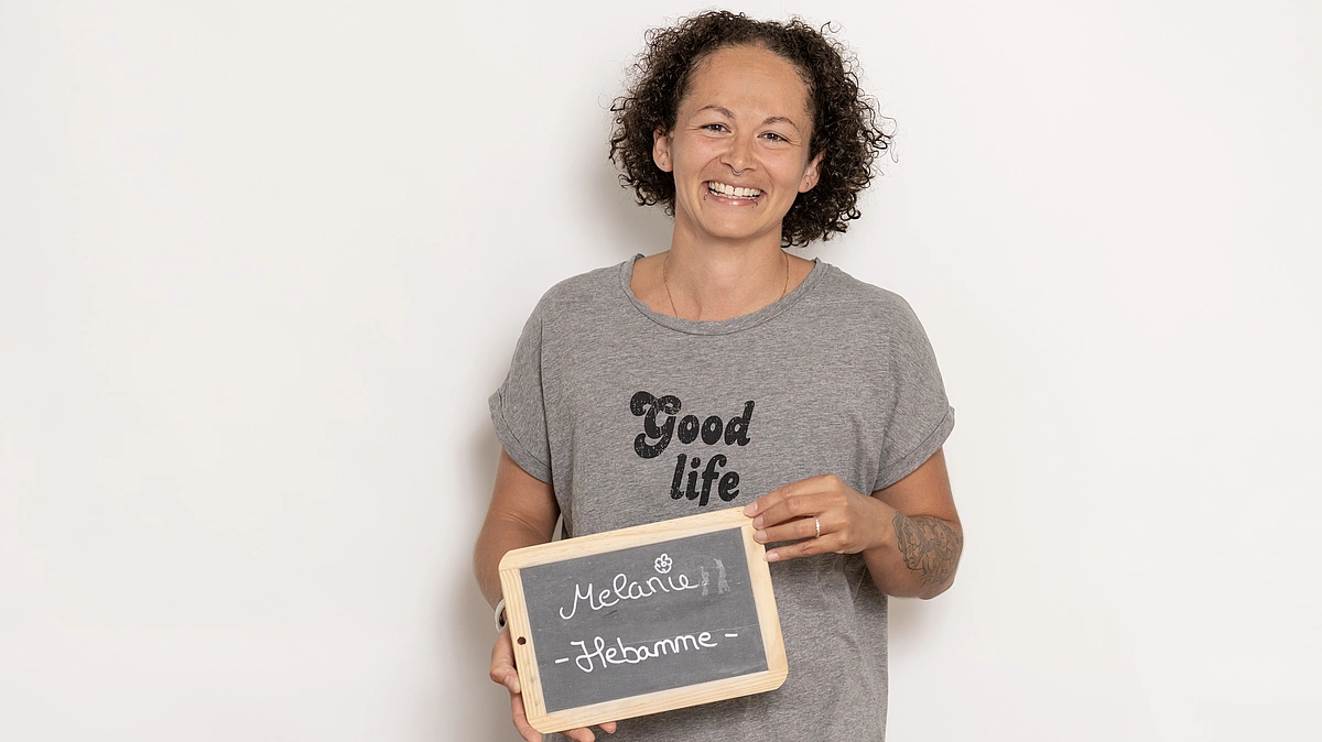 Hebamme Melanie der Hebammenpraxis Meilenstein zeigt mit einem großen Strahlen eine Tafel mit ihrer Berufsbezeichnung in Kirchheim unter Teck. Sie hat schwarze lockige Haare und trägt ein grau melierte T-Shirt mit schwarzem Aufdruck "Good life".