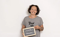 Hebamme Melanie der Hebammenpraxis Meilenstein zeigt mit einem großen Strahlen eine Tafel mit ihrer Berufsbezeichnung in Kirchheim unter Teck. Sie hat schwarze lockige Haare und trägt ein grau melierte T-Shirt mit schwarzem Aufdruck "Good life".