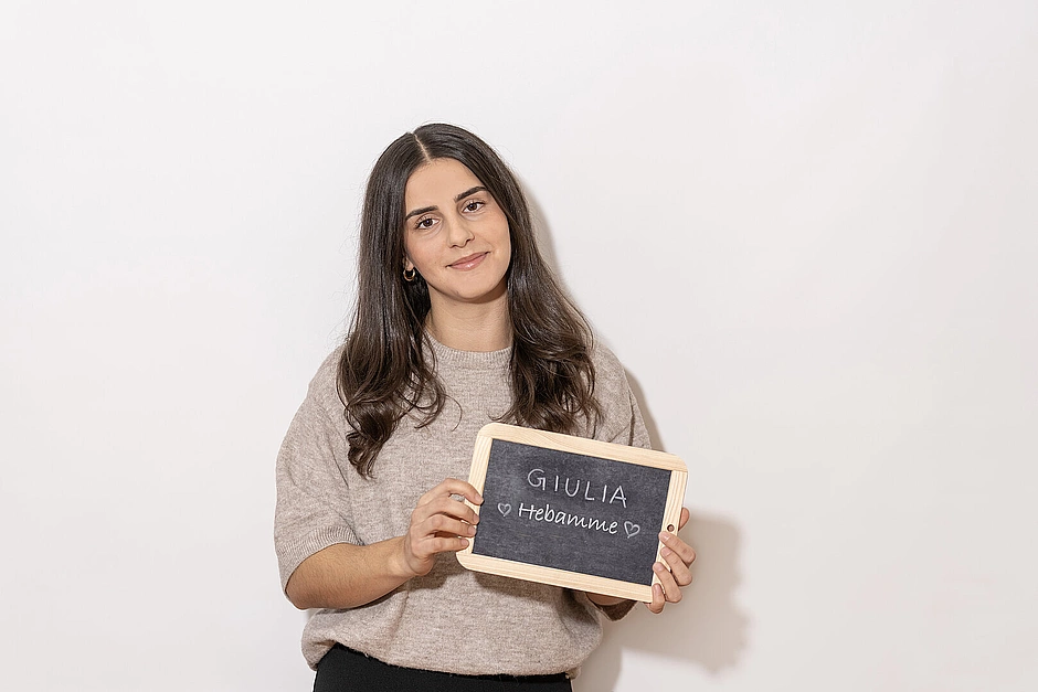 Frau in beigem Pullover hält eine kleine Kreidetafel mit der Aufschrift ‚Giulia – Hebamme‘ vor hellem Hintergrund.