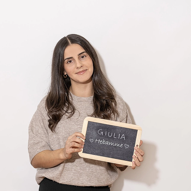Frau in beigem Pullover hält eine kleine Kreidetafel mit der Aufschrift ‚Giulia – Hebamme‘ vor hellem Hintergrund.