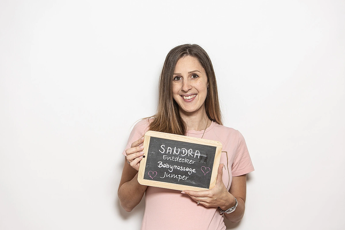 Frau in rosa T‑Shirt hält eine Kreidetafel mit der Aufschrift ‚Sandra – Entdecker, Babymassage, Jumper‘ vor weißem Hintergrund.
