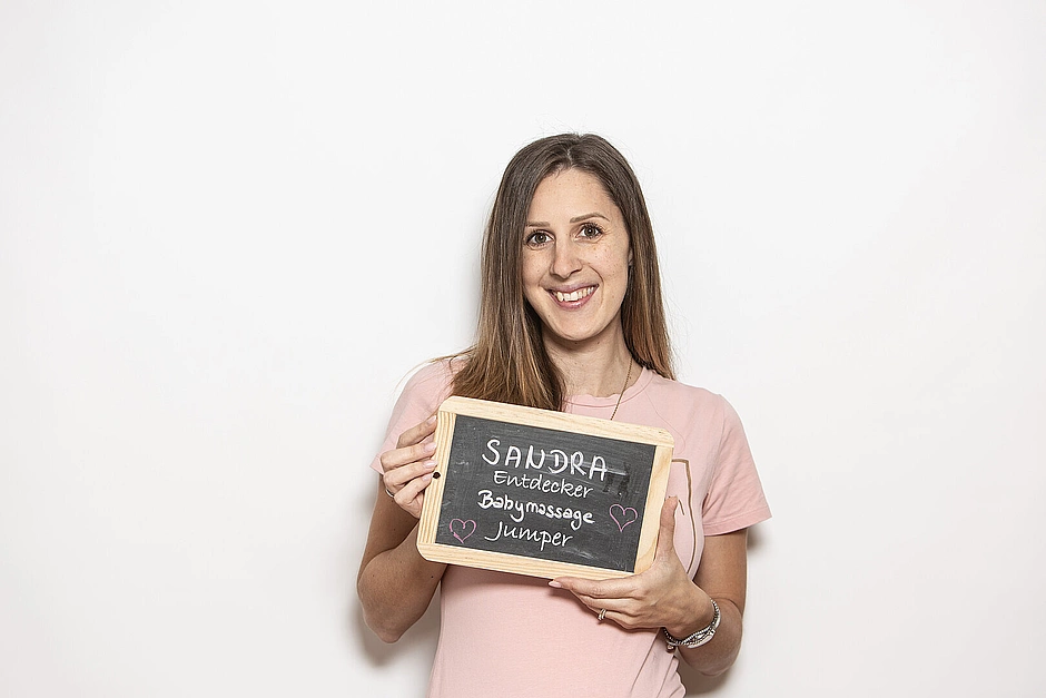 Frau in rosa T‑Shirt hält eine Kreidetafel mit der Aufschrift ‚Sandra – Entdecker, Babymassage, Jumper‘ vor weißem Hintergrund.