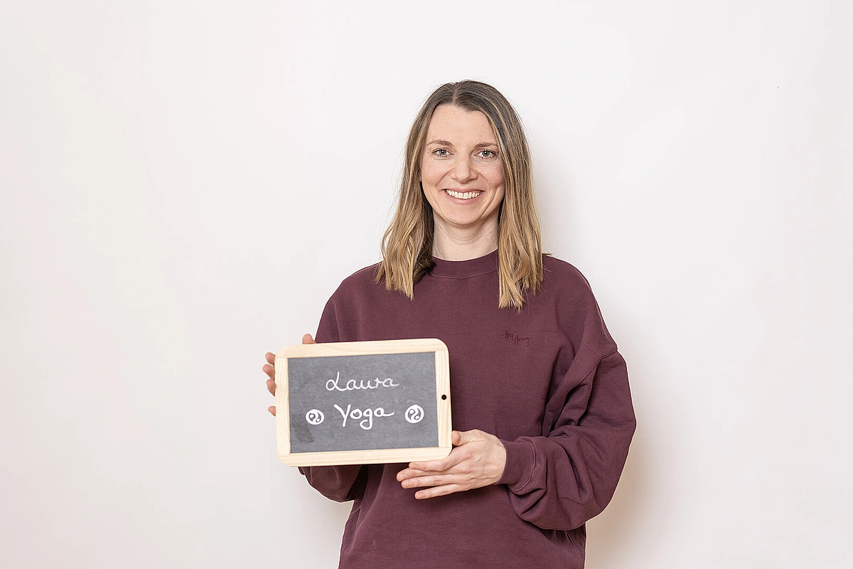 Frau in bordeauxfarbenem Pullover hält eine kleine Kreidetafel mit der Aufschrift ‚Laura – Yoga‘ vor hellem Hintergrund.