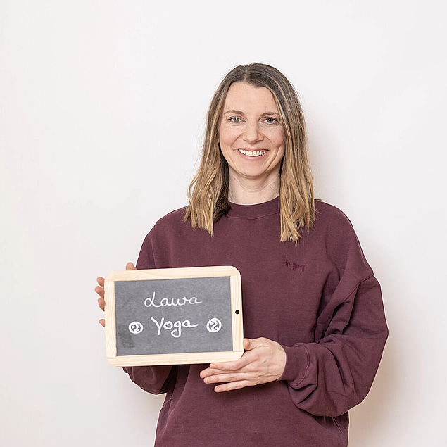 Frau in bordeauxfarbenem Pullover hält eine kleine Kreidetafel mit der Aufschrift ‚Laura – Yoga‘ vor hellem Hintergrund.